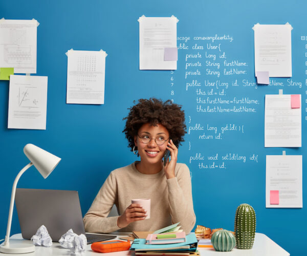Cheerful female student has coffee break, calls someone via cellphone, sits at working place near laptop computer, shares good news about passing exam successfully, uses free internet connection