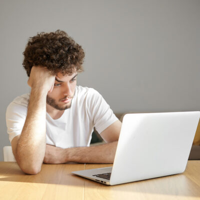 Horizontal shot of frustrated young unshaven male freelancer in white t-shirt using electronic device for distant work, having tired or bored look while working on urgent project early in the morning